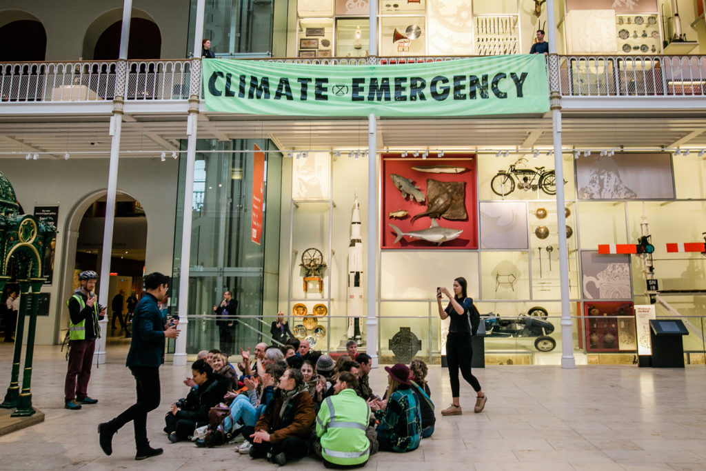 Protesters sit on floor in middle of museum as other protesters hang flag from balcony saying Climate Emergency