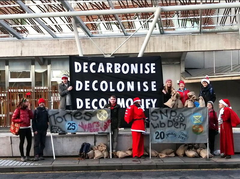 Rebels stand in front of a banner saying Decarbonise, Decolonise, Decomodify