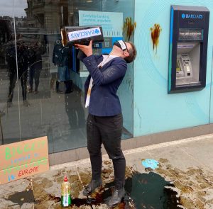 Climate activists stage performance outside a Barcalays bank. Person dressed as Barclays worker about to pour oil on themselves.