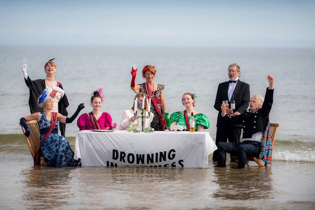 Rising sea engulfs Edwardian tea party on the beach. Banner says Drowning in Promises.