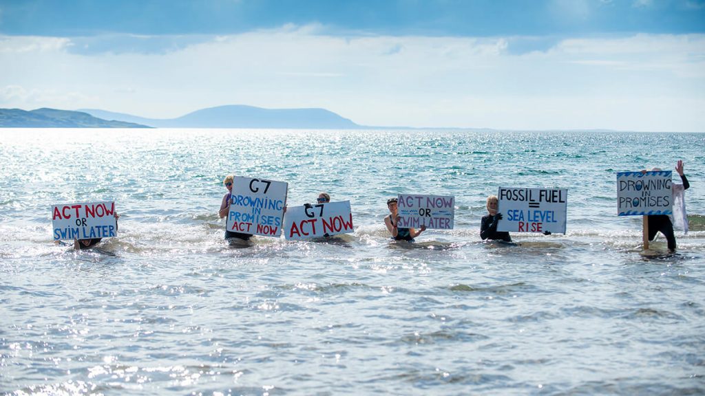 Activists standing in sea with placards saying 'Act Now or Swim Later' or 'Fossil Fuel = Sea Level Rise'