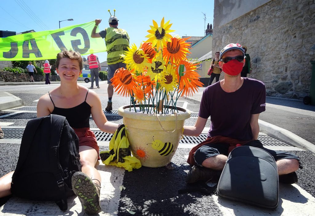 Two people sitting on road locked onto pot with colourful artificial flowers.