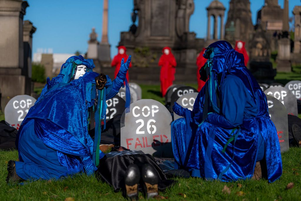 Blue Rebels grieve around a dead person acting lying on ground in front of sign saying COP26 Failed. In background Red Rebels and tombs of Glasgow Necropolis.