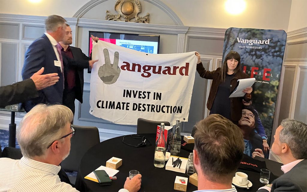 Two climate activists hold up a banner in a Vanguard meeting saying 'Vanguard invest in climate destruction'. A man tries to pull the banner down.