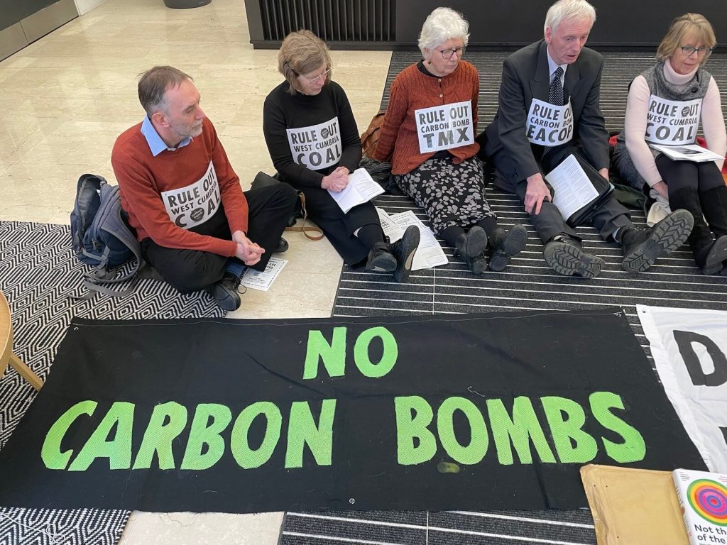 XR Scotland climate activists sit on floor of insurance company AIG. They sit around a flag that says 'No Carbon Bombs'. They wear messages that says Rule ut West Cumbria Coal, Rule out Carbon Bomb TMX and Rule out Carbon Bomb EACOP