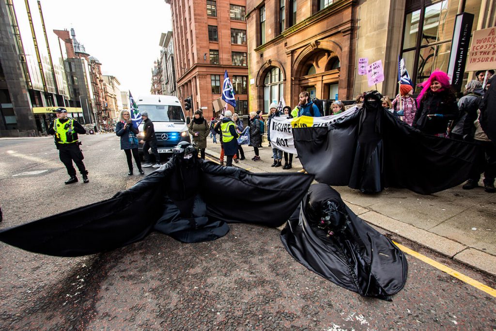 Oil Slicks, a protest troupe dressed head to toe in black, perform in front of protest with one kneeling on road an danother prostrate on road. Activists wave placards and XR flags.