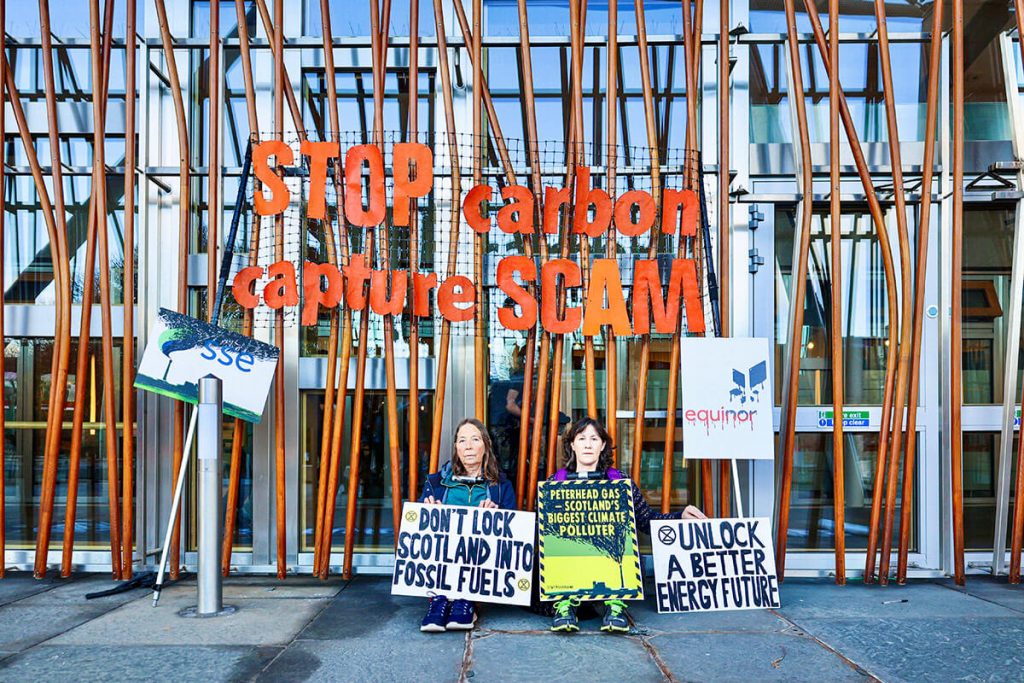 Two activists locked-on by their neck to the Scottish Parliament. They sit under a banner saying: 'Stop carbon capture scam'. One activist holds a banner saying 'Don't lock Scotland into fossil fuels'. The second activist holds a placard saying: 'Peterhead gas = Scotland's biggest climate polluter' and another placard saying 'Unlock a better energy future'.