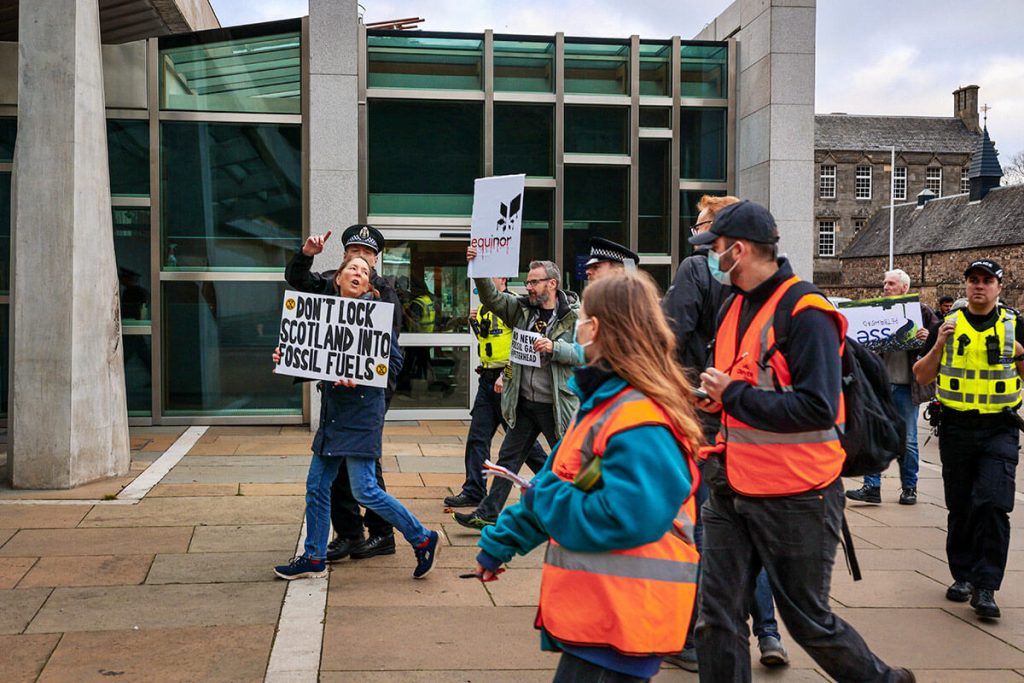 Arrested XR Scotland activist being led away by police outside Scottish Parliament during carbon capture is a scam protest. Arrested activist holds placard saying 'Don't lock Scotland into fossil fuels'.