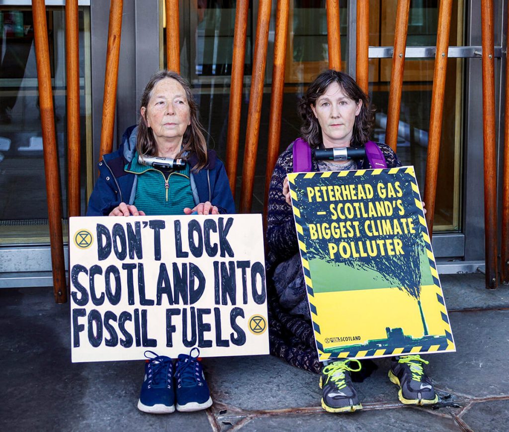 Two activists locked-on by their neck to the Scottish Parliament. They sit under a banner saying: 'Stop carbon capture scam'. One activist holds a banner saying 'Don't lock Scotland into fossil fuels'. The second activist holds a placard saying: 'Peterhead gas = Scotland's biggest climate polluter'.