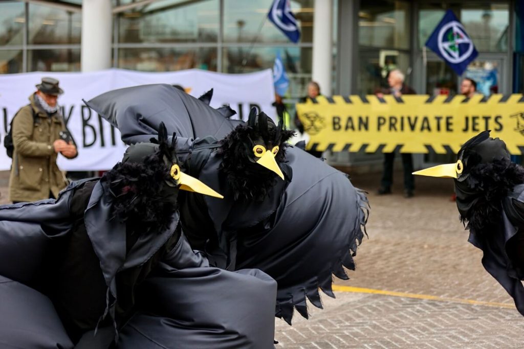 Protestors in blackbird costumes parade outside airport in front of banner saying Ban Private Jets.