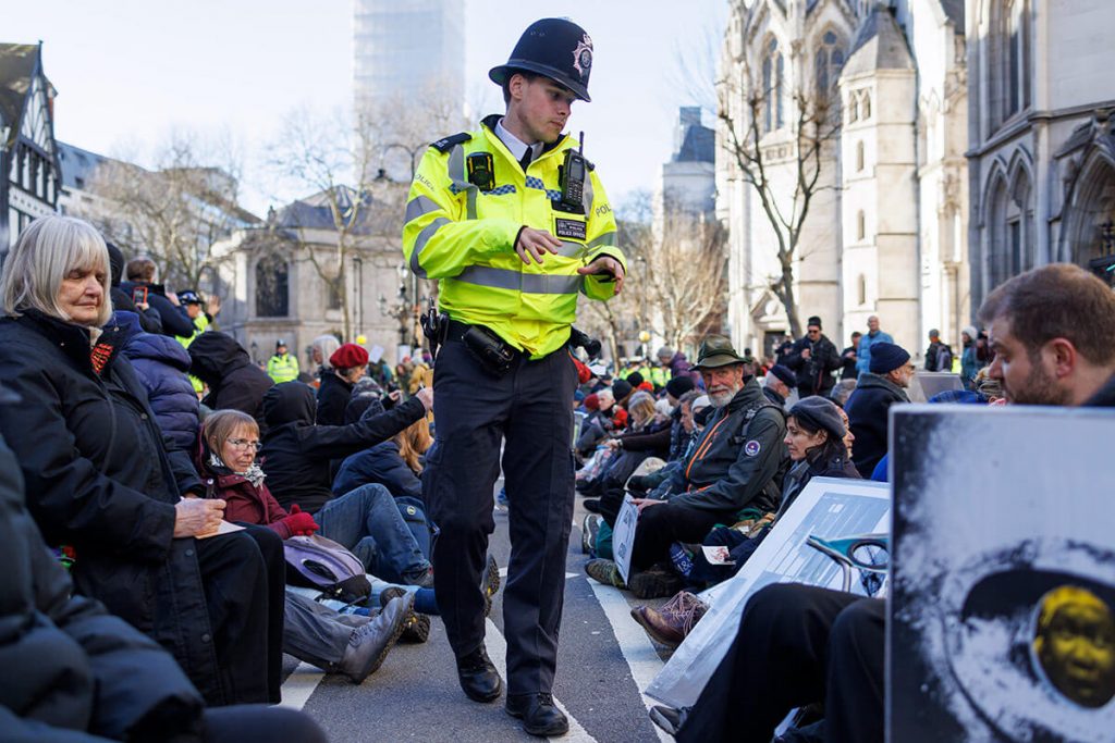 Policeman walks between two rows of protestors sitting down in road (the Strand outside Royal Courts of Justice, London). The activists are ignoring the police man as he talks to them.