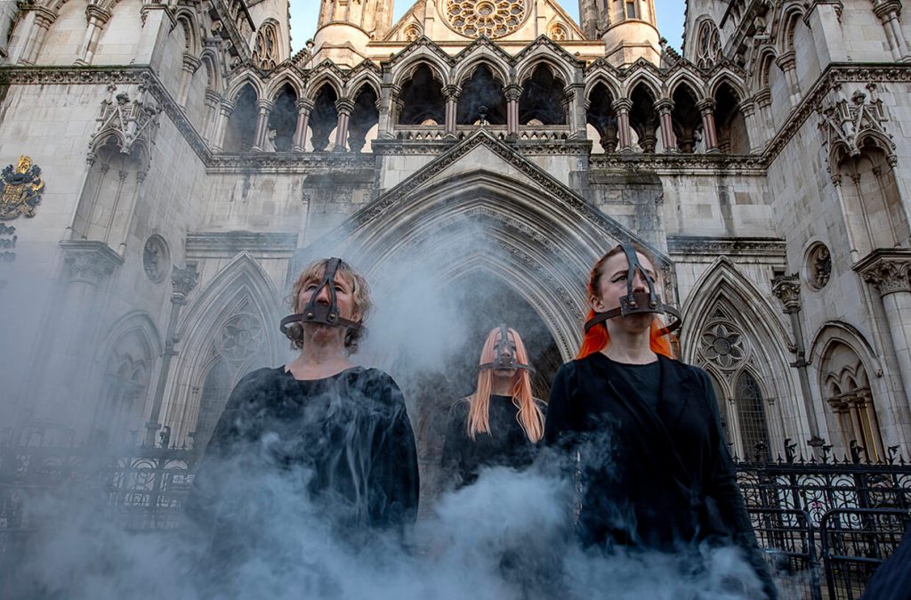 Three women wear scold’s bridles outside Royal Courts of Justice. They are dressed in black and smoke rises around them.