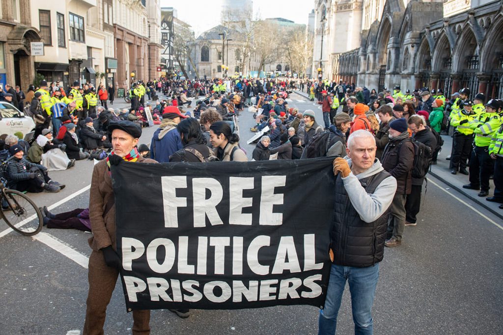 Two activists in foreground hold banner saying: Free Political Prisoners. Behind them, protestors sit down in road (the Strand outside Royal Courts of Justice, London). Groups of police stand next to the protest.