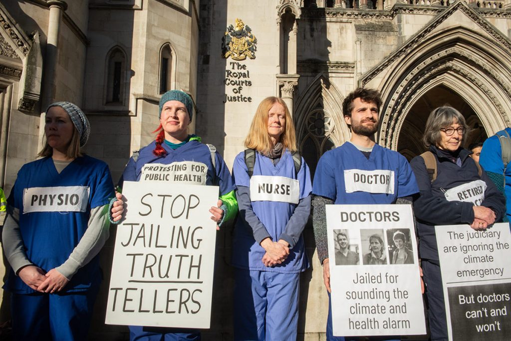 Healthcare workers in work uniforms stand outside Royal Courts of Justice. They hold signs saying 'Stop Jailing Truth Tellers' and Doctors - Jailed for sounding the climate and health alarm.